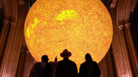 A large sun sculpture with a bright yellow and orange glow is suspended above a church-like building. The silhouettes of three people can be seen looking up at it, with one person pointing at it.