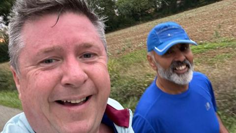 The picture shows two men in running in sports wear. One is red in the face wearing light blue and burgundy. The other is wearing a dark blue shirt and hat. 
