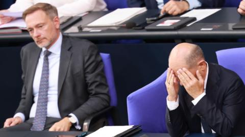 Finance Minister Christian Lindner and Chancellor Olaf Scholz sat together. Lindner is wearing a suit and Scholz has his head in his hands. 