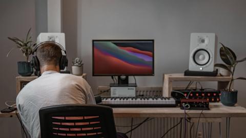 A man sits with his back to the camera at a music production desk. He is to the left of the image, wearing a white cotton long sleeved t shirt, with short dark blonde hair and wears black headphones. He is sat at the wooden desk, which has a large computer screen in the centre, with a red wavy wallpaper design on the screen, a small keyboard is underneath the screen and there are two white speakers and small plant pots framing the desk at either end.