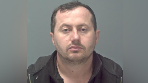 A police custody photo of Jani Murati, a man with short dark hair staring straight into the camera