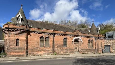 A general view of a red sandstone building with turrets, a dark roof and an arched white sign over a wooden door.