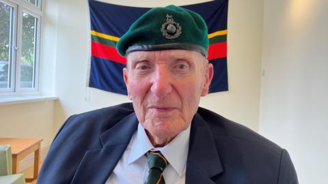 Tony Noakes looks directly at the camera. He is wearing a navy blazer, blue shirt, green striped tie and a green beret with a military emblem. 
