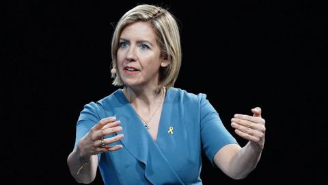 Dame Andrea Jenkyns giving a speech at a conference she has short blond hair and is wearing a blue dress and a headset microphone