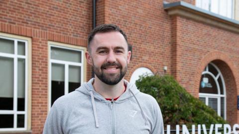 A man in a light grey zipped hoodie with a beard and short, dark hair is stood outside a red brick building. 
