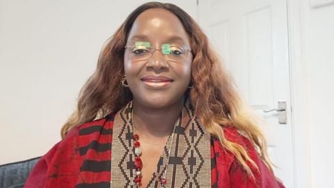Bami is wearing a red and black dress, which has black and cream patterned detailing around the neck-line. She has long, wavy light brown hair and is wearing frameless glasses. She is sat on a grey seat with a white wall and door behind her.