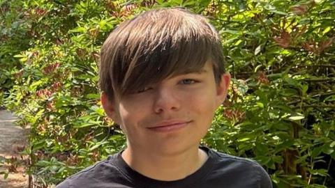 Joshua, wearing a dark T-Shirt and with his brown hair swept across his face, smiles for a photo in the sunshine