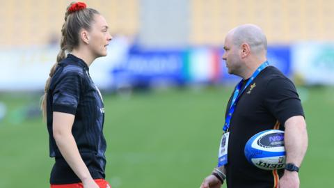 Hannah Jones in a training top talks to Sean Lynn who has a rugby ball under his arm on a pitch before a game.