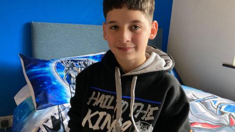 Teenage boy with dark hair wearing a Sheffield Steelkings hoody and sitting on his  bed which has blue, white and red Sheffield Steelkings bed covers