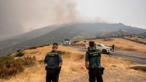 Two Civil guard officers look on as firefighters assemble with plume of wildfire smoke in the background in Avila, Spain on Monday