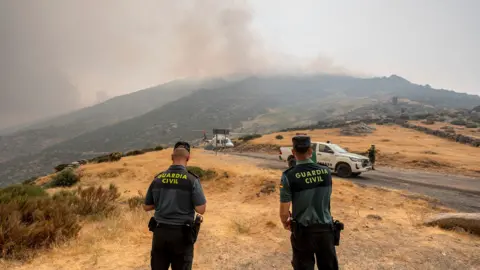 Two Civil guard officers look on as firefighters assemble with plume of wildfire smoke in the background in Avila, Spain on Monday