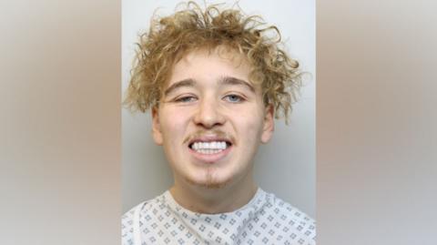 Teddy Kelt smiles in his police custody photo. He has blonde curly hair. He is wearing a hospital gown. 
