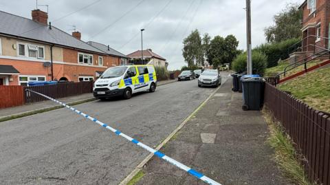 A police van is parked in a residential street with police cordon tape blocking the road. Houses and bins are pictured on either side of the road