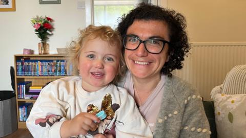 Georgie Murray-Gourlay is holding her daughter Lilly in her arms. Georgie has dark curly hair and Lilly has blonde curls. They are both smiling. They are sitting at home on a sofa, with a bookcase in the background.