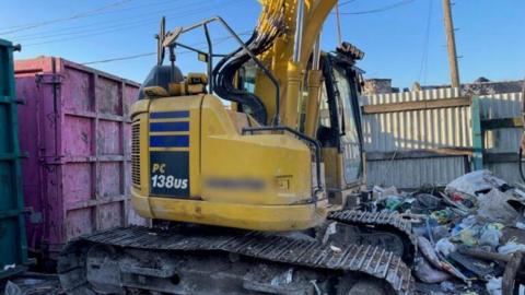 A yellow excavator with a cab on a floating platform in a waste and recycling site next to a pink shipping container and behind a fence