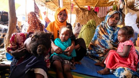 Two women sit holding two young children. An older child sits to the left of them with her face in her hands. They are in a shelter in a displacement camp after fleeing El-Fasher.