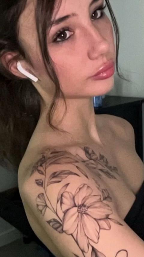 Levi is taking a selfie. She has long dark brown hair with two strands of hair loose to frame her face. She has a white wireless headphone in her ear and a sleeve tattoo from her shoulder down her arm of a fine flower art piece.