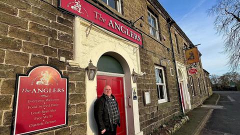 Rick Ellison stood outside the Anglers in front of a red door, with a sign saying The Anglers above the door. 