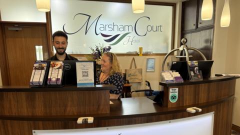 A woman and a man stood at a desk in a hotel looking at a computer screen.