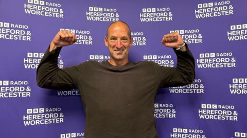 Mark Bond pictured smiling with his arms flexing his muscles by his side. He is wearing a long sleeve brown jumper and is bald.