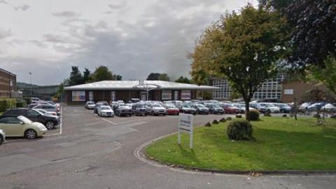 Tiverton High School with an entrance sign at the front on some green grass, with a car park full of cars in front of the building.