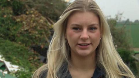 A girl with flowing blonde hair is wearing a black top and talking to the camera. She is stood in a field.