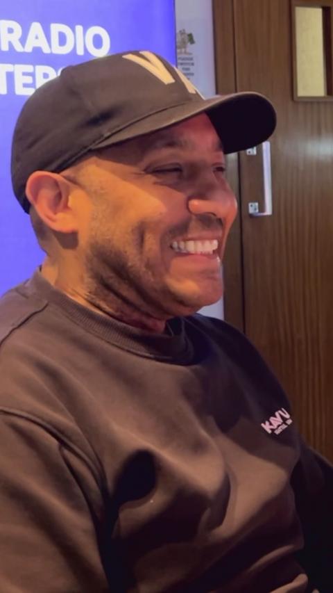 Man sitting in radio studio wearing brown jumper and hat.