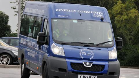 A dark blue Beverley Community Lift minibus being driven past a row of cars. The driver is visible through the windscreen and can be seen behind the steering wheel wearing a high-vis jacket.