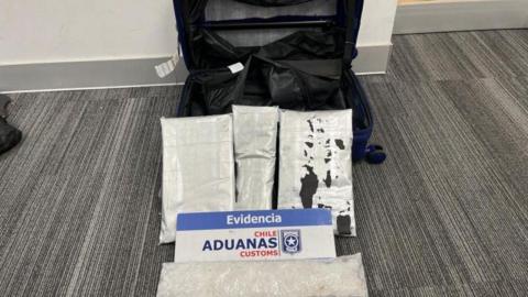 A cabin-sized suitcase which has been cut to reveal a secret compartment pictured next to bags of white powder which is methamphetamine. There is a sign by the bags of powder which says 'Evidencia Chile Aduanas Customs'.