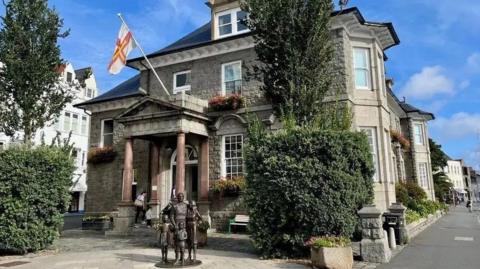 The exterior of the States Offices on a blue sky day. There are sculptures of a man, woman and two children in the square in front of the building. There is a tree, bush and granite planter of flowers either side of the entrance. The entrance has a portico with columns which has the Guernsey flag flying from it. The building has three storeys and stands next to a road.
