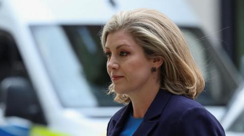 Penny Mordaunt is pictured walking outdoors. She wears a dark suit. 