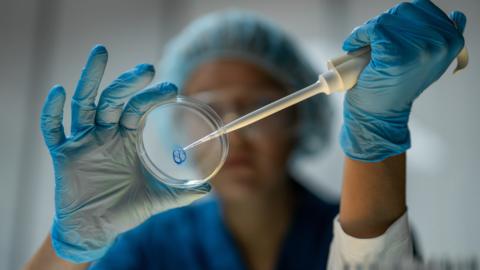 A female lab technician wearing full PPE uses a pipette to place a drop of fluid in a petri dish.