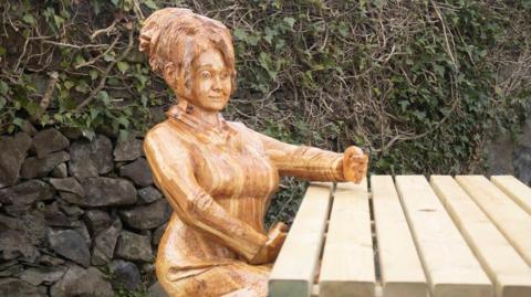 A wooden sculpture of a woman sitting at a picnic bench