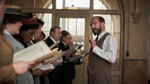 A group of people in historical costume stand in a line, holding sheet music, mid-rehearsal. A man, Ralph Fiennes, in a white shirt and waistcoat gestures with one hand, likely conducting.
