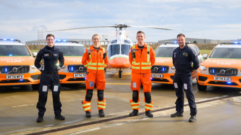 Team of Magpas air ambulance crew- three men and a woman- with two of them wearing orange uniform and the other two wearing black uniform, standing in a semi-circle in front of orange emergency vehicles and a helicopter. 