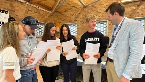 A male and a female teacher either side of four pupils who are holding their results