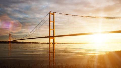 The sun sets behind the Humber Bridge. The sun's rays shimmer on the calm water. The reflections of both the north and south towers can be seen on the water.