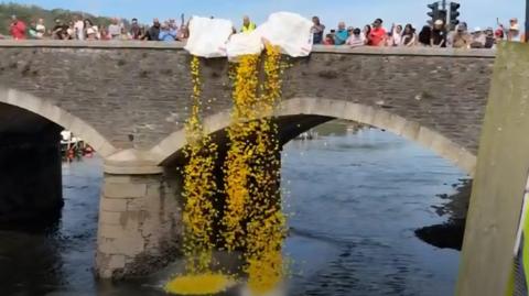 People on the bridge emptying bags of ducks. There are three people emptying the bags while other people along the bridge watch on. The bridge has three arches and there is a river below.