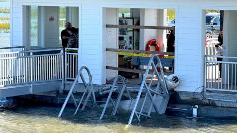 A close up photo shows the collapsed gangway on the Georgia dock, in front of one of the buildings on the dock, with police tape cordoning off the area and a few people looking on.