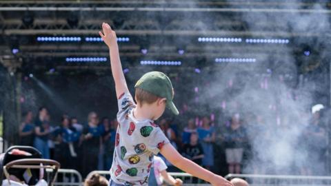 A child wearing a grey T-shirt with superheroes on it and a green cap dancing with his hands in the air. He is facing a stage with performers on and dry ice smoke.