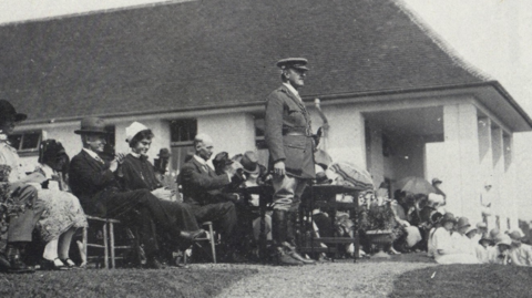 A picture of Field Marshal Viscount Allenby standing to address people gathered outside the hospital at its official opening, with seated dignitaries gathered behind him. 
