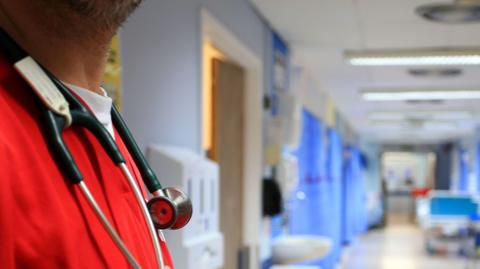 A stock image of a man's upper body, cropping out his face, showing him wearing a stethoscope and red scrubs. In the background is a blurred hospital ward.