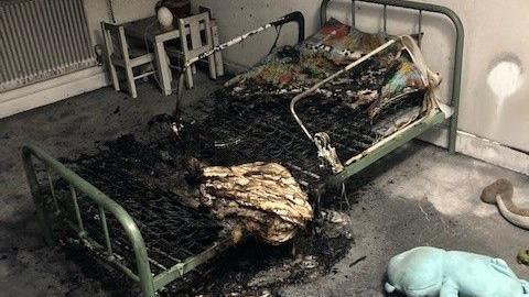 A bed completely destroyed by a fire with cuddly toys on the carpet