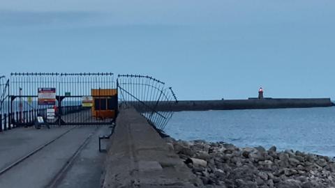 The pier which is a concrete structure which curves to the right. There is a small lighthouse towards the end which is lit up. There are metal gates with spikes at the top. There are various signs on the gate. It is dusk and the sky is a grey-blue colour and the sea beside the pier is calm.