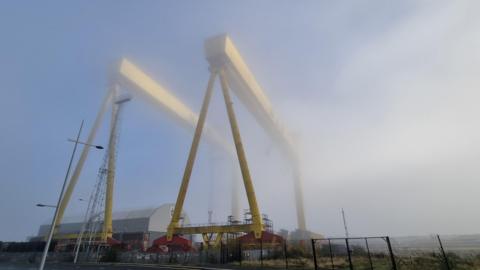 Industrial cranes and sweeping fog. The two cranes are yellow. An industrial building is under the cranes.