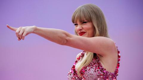 Taylor Swift points into the distance with a grin and her eyes squinted beneath her blonde fringe. She wears a sparkly pink fitted bodice and red lipstick. The background is pinkish-purple gradient and matches her outfit.