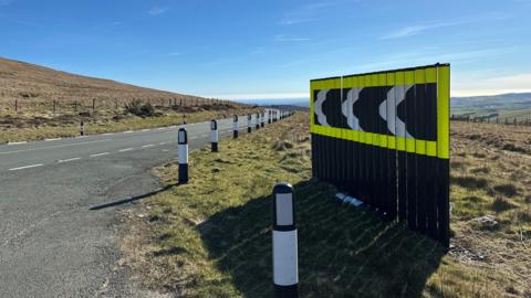 Large section of black and white chevrons framed in luminous yellow positioned at the side of the road at Brandywell. The road sweeps round a bend in the distance with blue sky overhead.