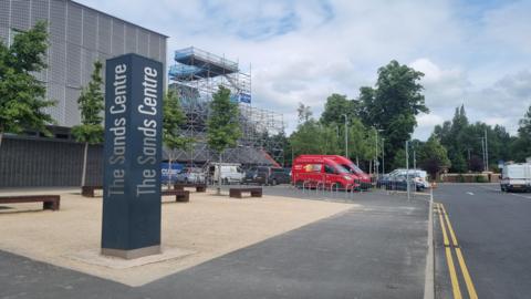 A general view of the entrance to The Sands Centre in Carlisle. A black pillar has the name of the venue written on each side in silver. The main building is behind the pillar and on the right there is an area of the building surrounded by scaffolding.