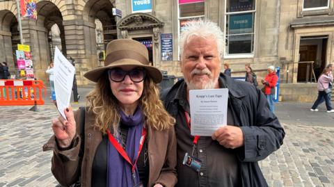 A woman in large sunglasses and a large-rimmed hat as standing beside a man with grey hair and beard in the street as they hold fliers for their play
