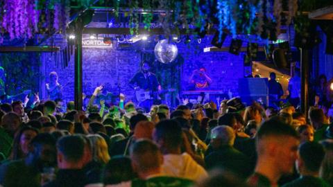 A crowd of people in a venue are seen with a band on stage. There is a decorative floral arrangement hanging from the ceiling and a disco ball. The room is illuminated in a blue light.